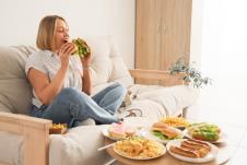 Žena sedi na dvosedu u ruci drži dva sendvič-burgera, a ispred nje na stolu se nalazi izobilje hrane