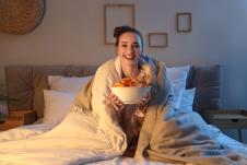 Devojka sedi u krevetu ogrnuta jorganom, a u rukama drži činiju čipsa