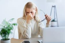 zena u bež džemperu sa zatvorenim očima sedi ispred računara i desnom rukom se drži za koren nosa a levom rukom drži naočare