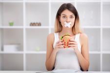 devojka sa flasterom preko usta sedi za kuhinjskim stolom i drži sendvič