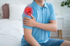 muškarac sa bolom u ramenu u plavoj majci sa kratkim rukavima sedi na ivici kreveta i drži levu šaku na desnoj nadlaktici