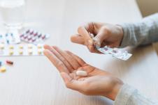 žena u levoj ruci drži tabletu, a u desnoj blister sa lekovima ispred nje na stolu je nekoliko blistera sa različitim tabletama i kapsulama