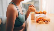 žena s viškom kilograma drži burger, ispred nje na stolu brza hrana