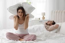 Devojka sedi na krevetu sa jastukom na glavi dok muškarac leži na drugoj strani krveta na leđima i hrče