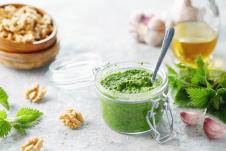 pesto od koprive serviran u maloj staklenoj tegli na stolu pored koprive, belog luka i oraha