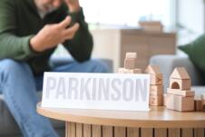 Papir sa rečju parkinson i drvene kocke na stolu, u pozadini čovek sa ukočenom rukom sedi kod kuće
