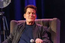 Čarli Šin sedi na fotelji, drži mikrofon i oblizuje se