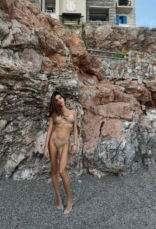 Dejana Živković stoji na šljunkovitoj plaži ispred visoke stene, u bež bikiniju