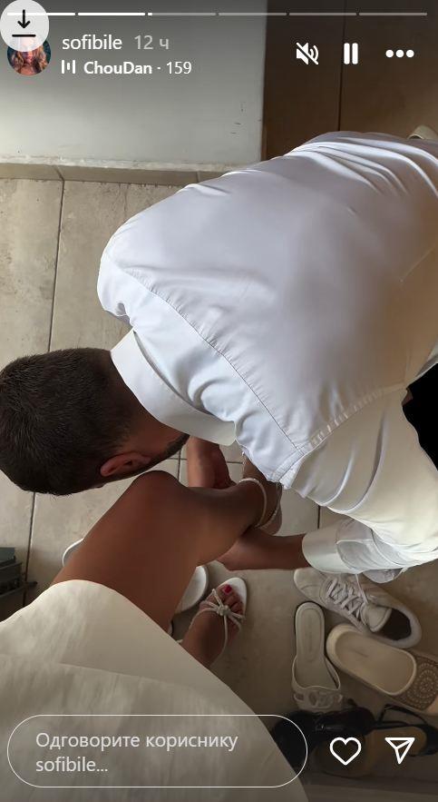 Sofi Tkačenko pravi selfi u ogledalu, dok muškarac u beloj košulji kleči ispred nje i oblači joj sandale