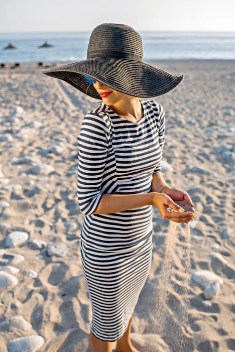 stockphotoyoungandelegantwomaninstrippeddresswithahatpouringsandwiththehandsonthebeachon289971479.jpg