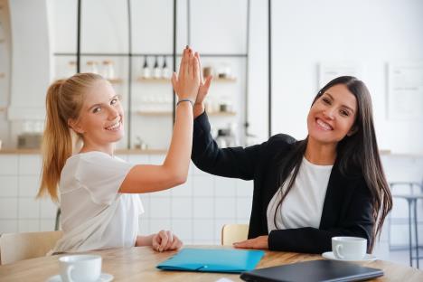 happyyoungbusinesswomengivinghighfivecelebratingsuccesssittingtablewithdocumentscoffeecupslookingcamera.jpg