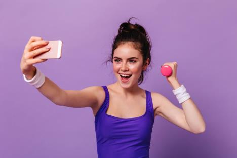 portraityoungwomanfitnesstopholdingpinkdumbbelltakingselfiepurplewall.jpg