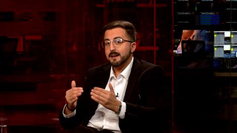 Čovek sa naočarima, u crnom sakou i beloj košulji u televizijskom studiju crvene boje