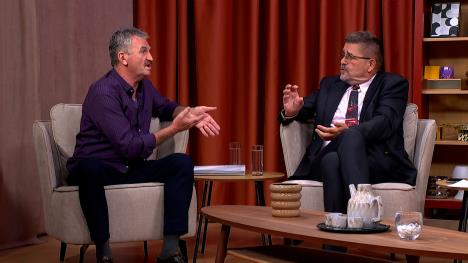 Dva starija muškarca sede ne bež foteljama u televizijskom studiju. Između njih dvojice je sto sa dve čaše vode