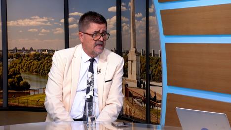 Muškarac sa naočarima u belom sakou i beloj košulji, sa kravatom u televizijskom studiju