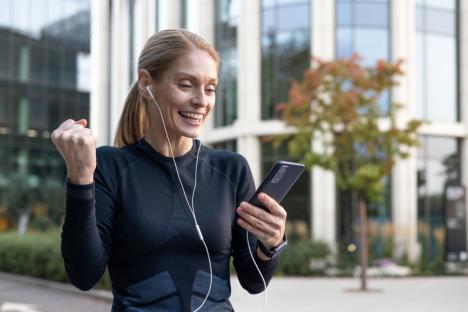 devojka sa plavom kosom vezanom u rep u crnoj sportskoj majci sa dugim rukavima i belim slušalicama drži telefon u levoj ruci dok je desna stisnuta u pesnicu