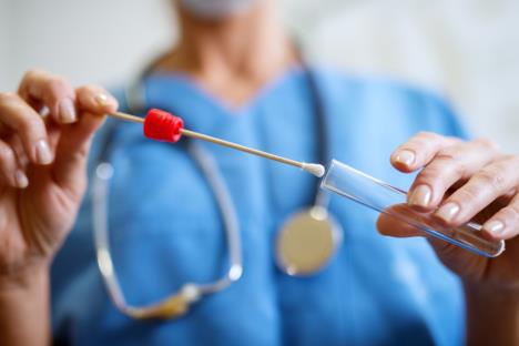 medicinski radnik u plavom odelu sa stetoskopom ubacuje u epruvetu štapić sa uzorkom za bris