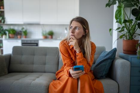 zabrinuta žena u narandžastoj haljini sedi na kauču s telefonom u ruci