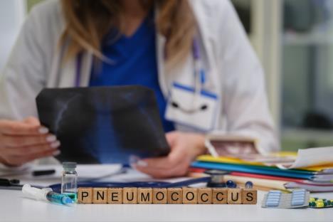 doktorka u belom mantilu sedi za sotlom i drži snimak pluća, na prednjem delu stola stoji natpis pneumomoccus na drvenim kockicama