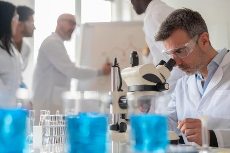 naučnik u belom mantilu u laboratoriji sa providnim zaštitnim naoračima gleda kroz mikroskop