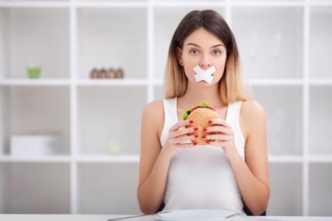 devojka sa flasterom preko usta sedi za kuhinjskim stolom i drži sendvič