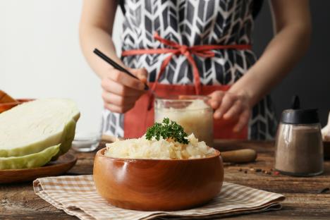 žena sa keceljom u kuhinji, na stolu kupus ribanac u drvenoj činiji, pola glavice kiselog kupusa, biber u teglici