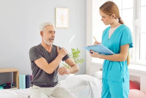 muškarac srednjih godina sedi na stolu za pregled kod lekara, drži se za levu ruku i objašnjava simptome doktorki