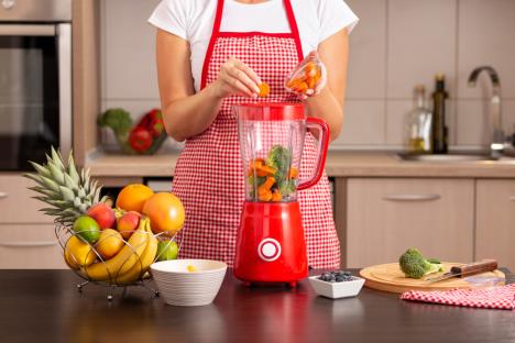 žena u crvenoj kecelji ubacje voće i povrće u crveni blender, koncept smuti