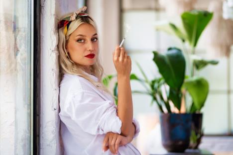 mlada žena se naslonila na zid i drži cigaretu u ruci, sobna biljka u pozadini