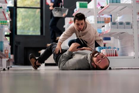 čovek koji ima napad epliepsije leži na podu, a pored njega je momak koji pokušava da mu pomogne