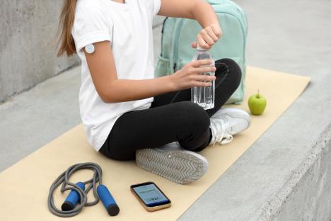 devojčica u crnim helankama i beloj majci sa aparatićem za praćenje nivoa šećera na nadlaktici sedi na podlozi za vežbanje pored vijače za preskakanje jabuke i otvara flašicu vode