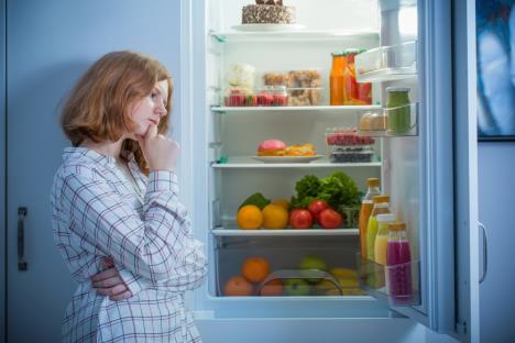 devojka u pidžami stoji ispred otvorenog frižidera i razmišlja da li da večera