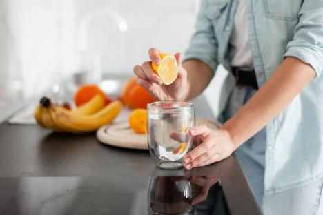 žena cedi limun u čašu vode, voće na stolu u pozadini