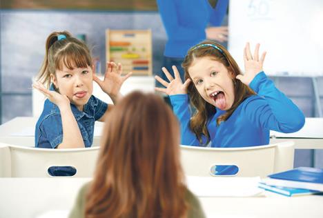 stockphotoelementaryageschoolgirlspullingfacestogirlsittingbehindteacheratblackboardin47236858.jpg