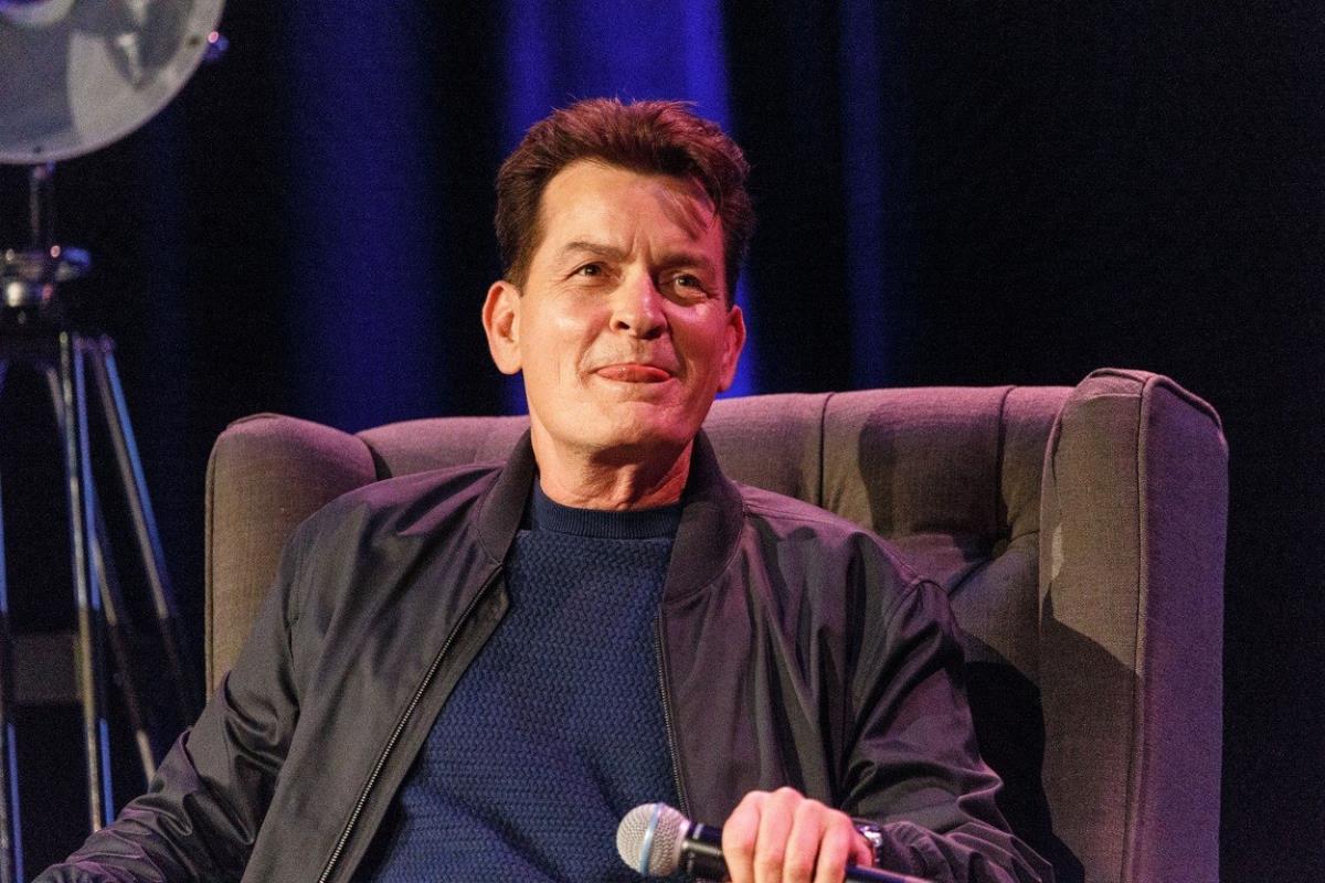 Čarli Šin sedi na fotelji, drži mikrofon i oblizuje se